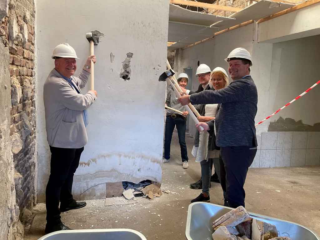 Foto 1: Weil das „Musterhaus“ in Nord-Düren nicht neu- sondern umgebaut wird, griffen Bürgermeister Frank Peter Ullrich (1.v.l.), Dr. Inken Tintemann vom Amt für Stadtentwicklung der Stadt Düren (2.v.l.) und der Technische Beigeordnete der Stadt Düren Niels-Christian Schaffert (3.v.l.) sowie Vertreter der Ratsparteien zum Start des Projekts zum Vorschlaghammer und nicht zum Spaten.