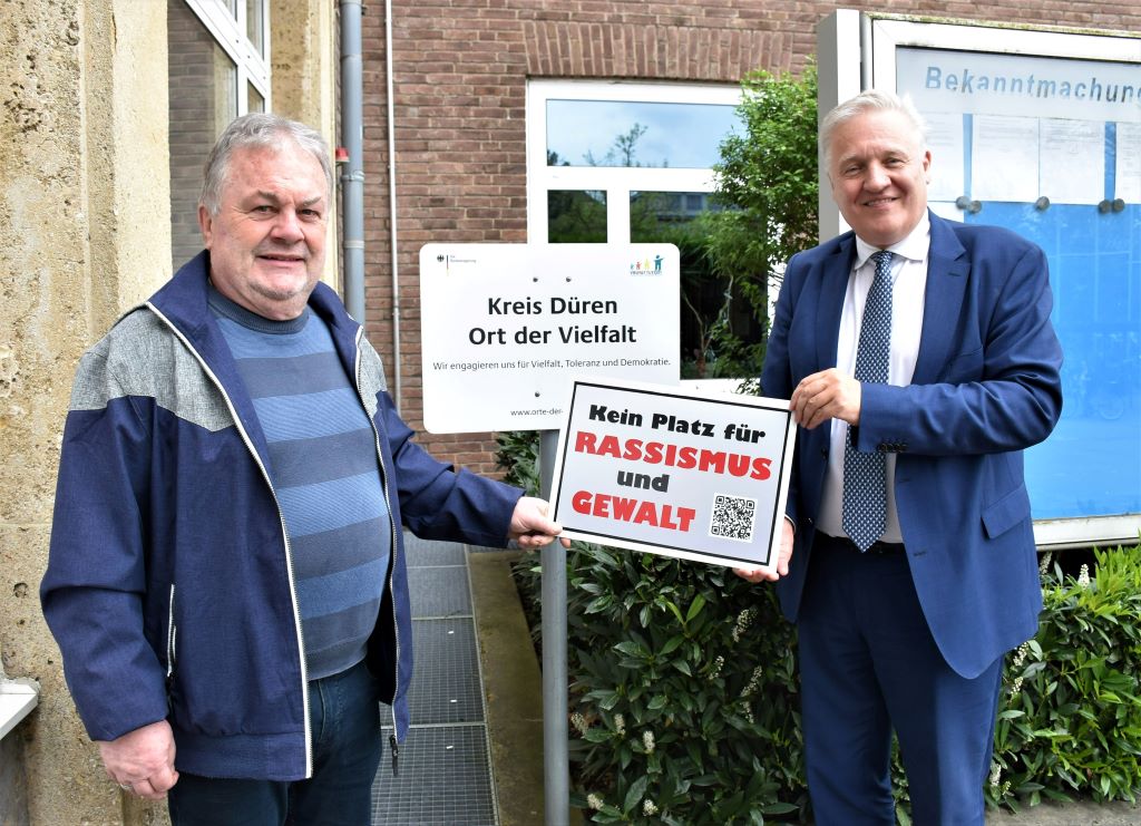 Joachim "Jo" Ecker (l.) hat Landrat Wolfgang Spelthahn eines seiner Schilder mit der Aufschrift "Kein Platz für Rassismus und Gewalt" überreicht. Es wird als klares Zeichen gegen Rassismus im Kreishaus aufgehangen. Foto: Kreis Düren. 